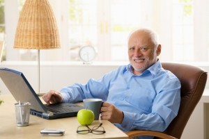 Portrait of happy senior man sitting at desk using laptop computer at home, smiling at camera.